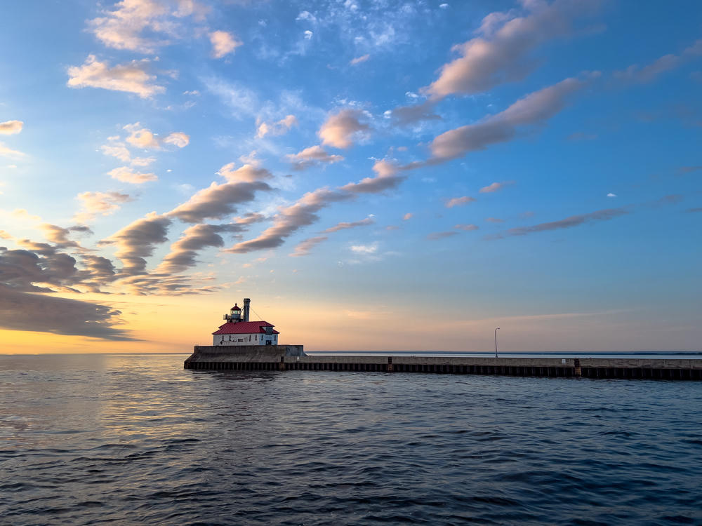 A white lighthouse with a red roof sits at the end of a long concrete and steel breakwater pier extending into calm Lake Superior at dawn, beneath a wide sky streaked with rows of golden and gray clouds.