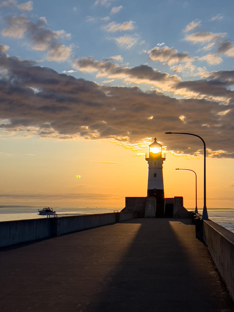 A concrete pier stretches toward a white lighthouse silhouetted against a glowing orange sunrise sky, with the sun blazing through the lighthouse’s lantern room. A small boat travels on the calm water to the left, and street lamps line the pier under a sky streaked with clouds.
