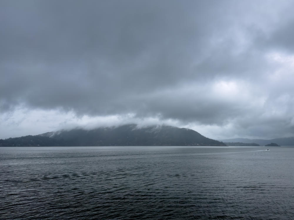 A forested island with its ridge shrouded in low cloud rises across a calm gray sea beneath a heavily overcast sky, with a small boat leaving a wake near the far right shoreline.