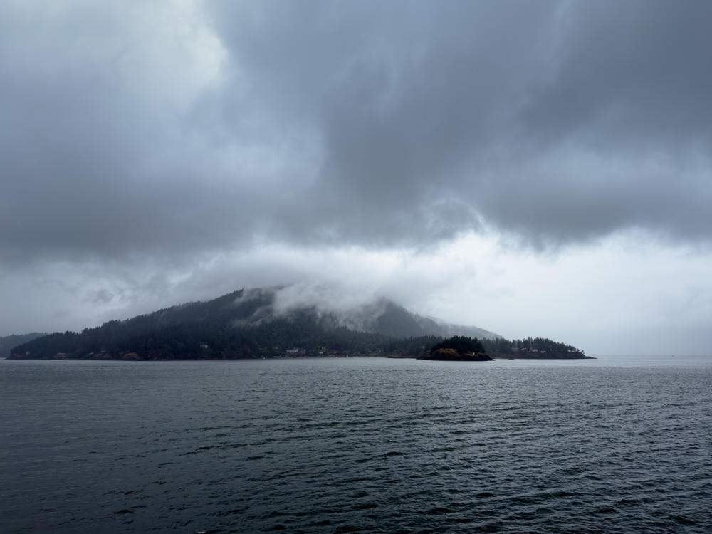 A moody, overcast view across grey rippled water toward a forested island whose hilltop is shrouded in low cloud and mist, with a scattering of waterfront houses along the shoreline near Bowen Island, British Columbia.