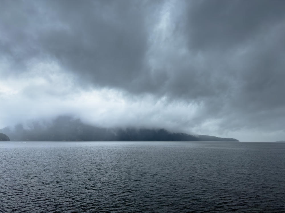 A moody, overcast seascape with low grey clouds and mist rolling over dark forested hills that meet a calm expanse of steel-grey water, seen from a ferry along the British Columbia coast.