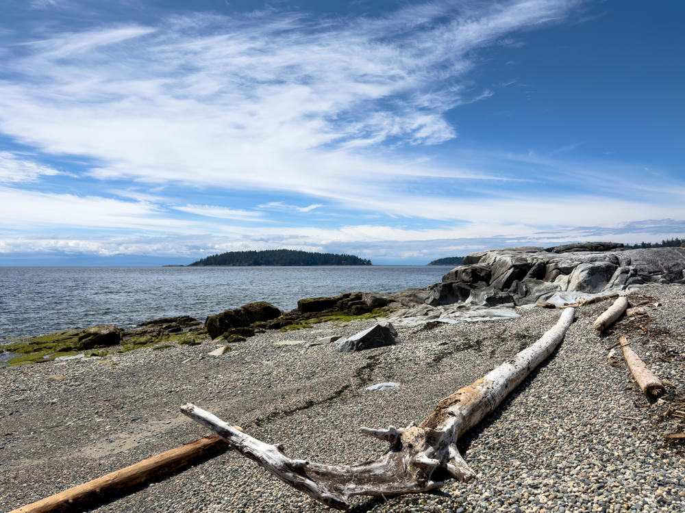 A rocky pebble beach with weathered white driftwood logs in the foreground, smooth grey rock formations to the right, and calm ocean water leading to a forested island under a bright blue sky streaked with wispy cirrus clouds.