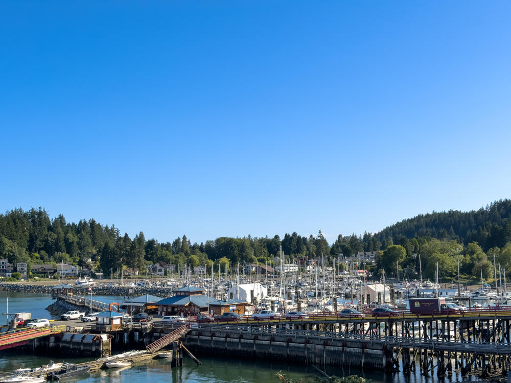 A busy harbour marina in Gibsons, British Columbia, packed with sailboats and pleasure craft, with a long wooden pier, waterfront buildings, and a parking lot full of cars in the foreground. Forested hills and scattered houses line the shore under a clear blue sky.