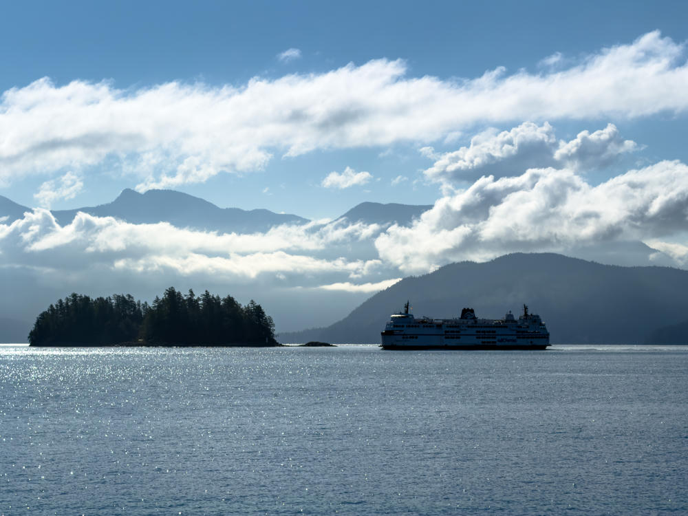 A BC Ferries vessel crosses shimmering blue coastal water past a small tree-covered island, with cloud-draped mountains rising in the hazy morning distance.