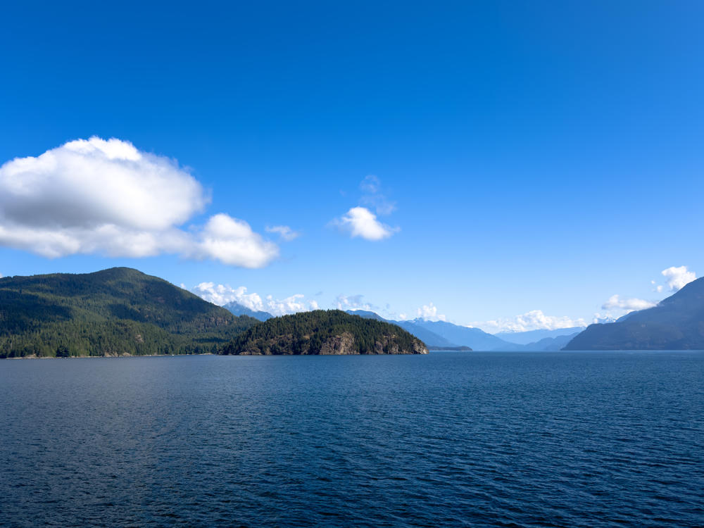 Forested hills and headlands rise from deep blue water along a coastal fjord, with layered mountain ranges receding into the distance under a bright blue sky dotted with white cumulus clouds.