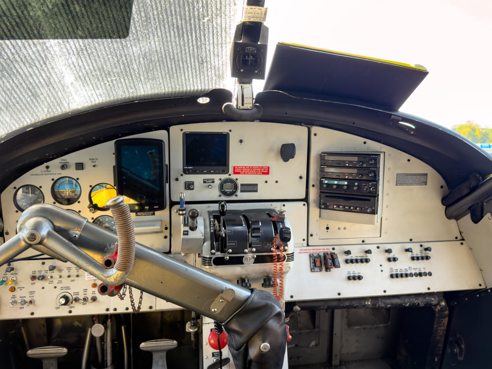 The cockpit of a floatplane with an extensive instrument panel of analog gauges, digital displays, radios, switches, and a control yoke, labeled “C-FHAA Harbour Express 309,” with a sun-shade over the left windshield and calm water visible through the right window.