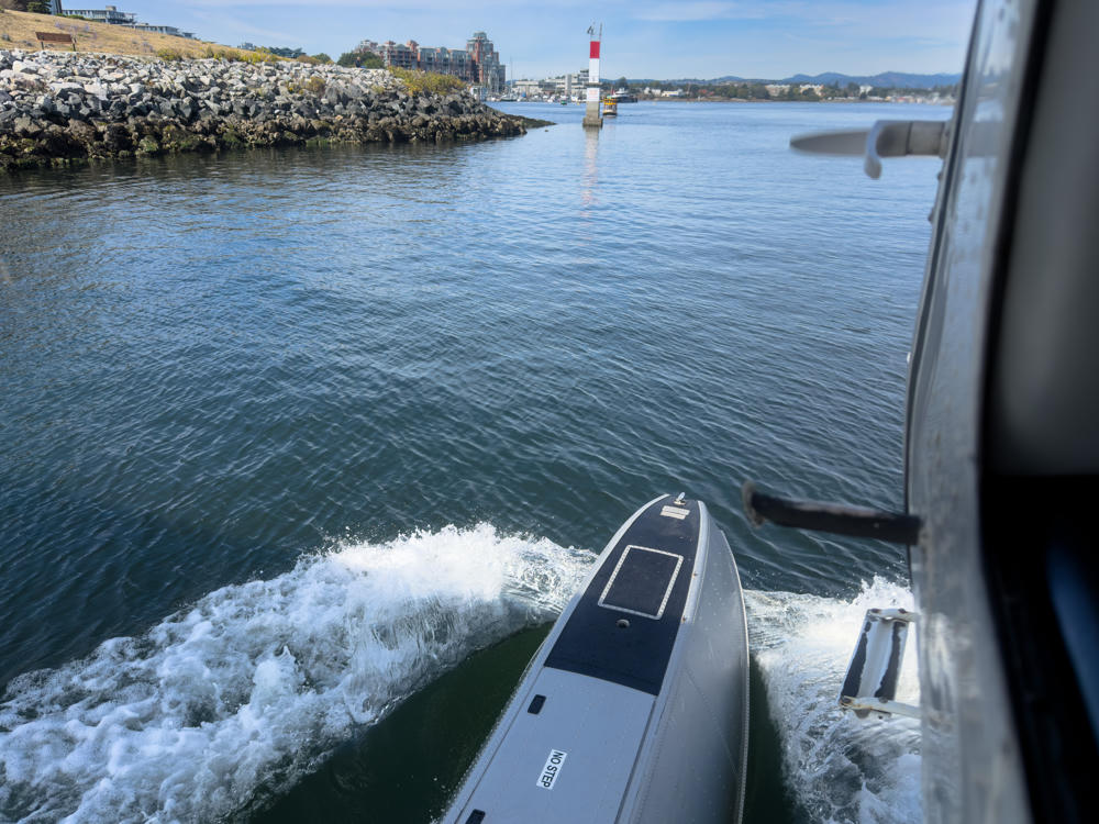 View from a moving seaplane’s float as it taxis across Victoria Inner Harbour, with churning white water spray beside the pontoon. A rocky breakwater with waterfront buildings lines the shore, a red-and-white channel marker beacon stands in the harbour, and distant hills rise under a partly cloudy blue sky.