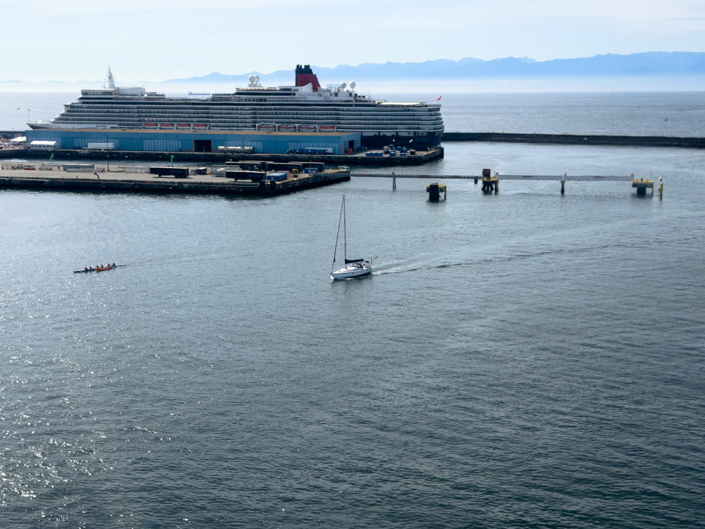 A large Cunard cruise ship with its distinctive red and black funnel docked at a harbour terminal, while a white sailboat and a small rowing shell move across the calm harbour water in the foreground, with distant hazy mountains along the horizon.