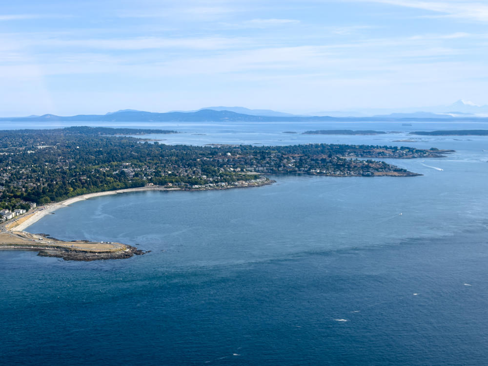An aerial view over the deep blue Salish Sea looking toward a wooded coastline dotted with houses, a curving sandy beach, and a low rocky point, with scattered islands and distant mountains including a snow-capped peak on the hazy horizon under a pale blue sky.