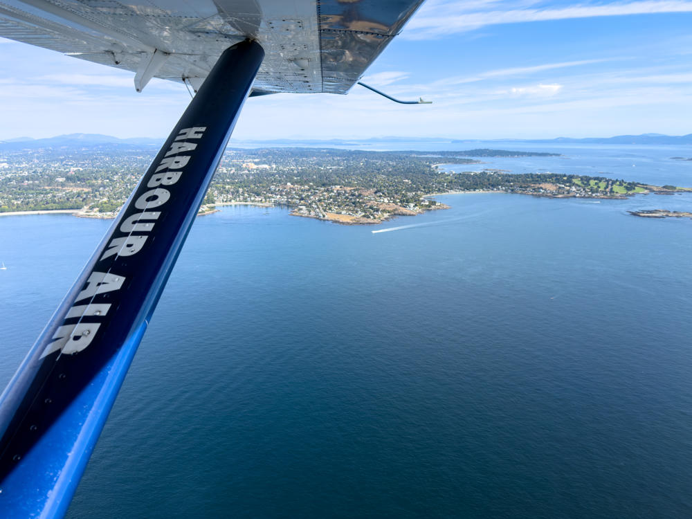 An aerial view from a Harbour Air seaplane, with the aircraft’s blue wing strut labeled “HARBOUR AIR” in the foreground, looking down over the blue waters of the Salish Sea toward a green, tree-lined coastline dotted with houses near Oak Bay, British Columbia, with distant islands and hills under a bright sky.