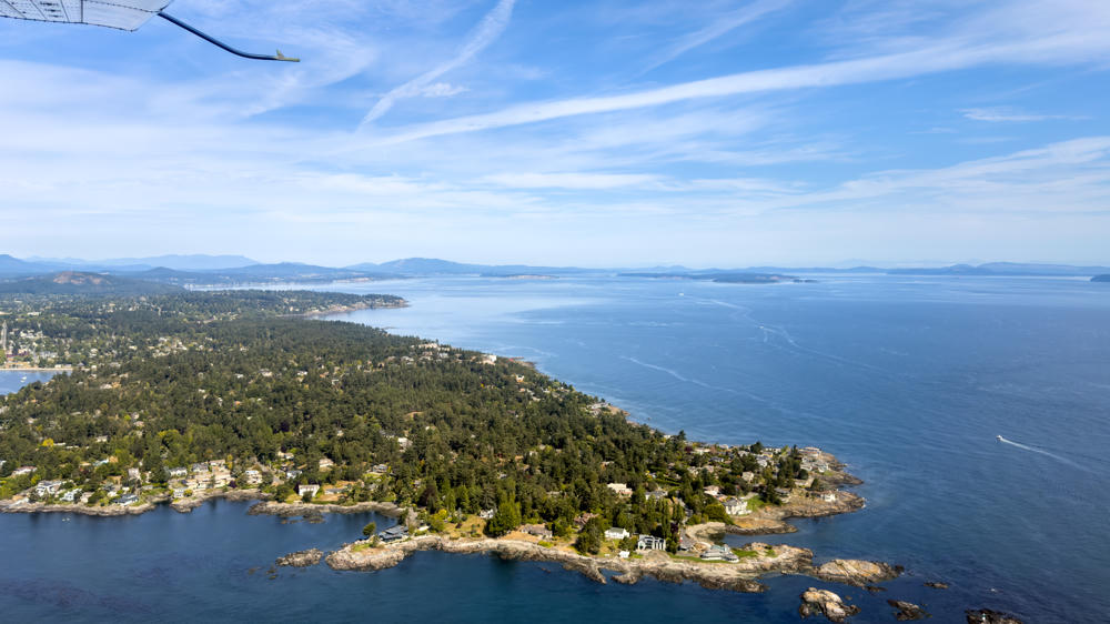 Aerial view from a small floatplane, with its wing strut visible in the upper left, looking down over a forested, rocky peninsula dotted with houses that juts into the deep blue Salish Sea. Distant islands and hazy mountains line the horizon under a bright sky streaked with wispy clouds.