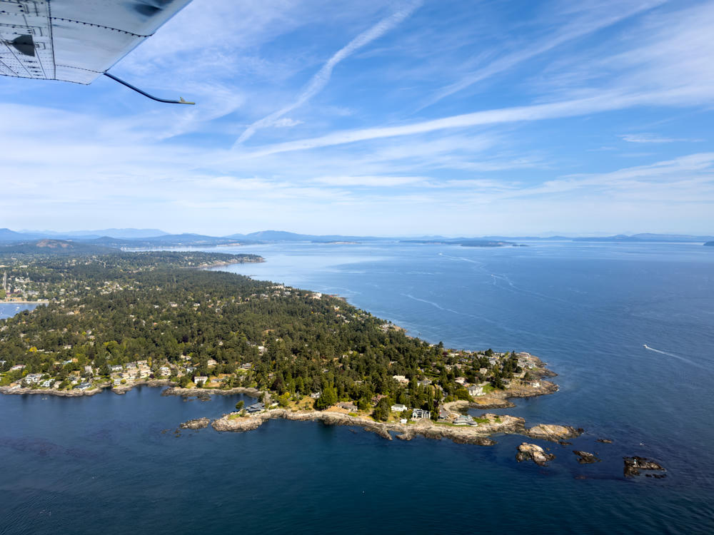 Aerial view from a small floatplane, with its wing strut visible in the upper left, looking down over a forested, rocky peninsula dotted with houses that juts into the deep blue Salish Sea. Distant islands and hazy mountains line the horizon under a bright sky streaked with wispy clouds.