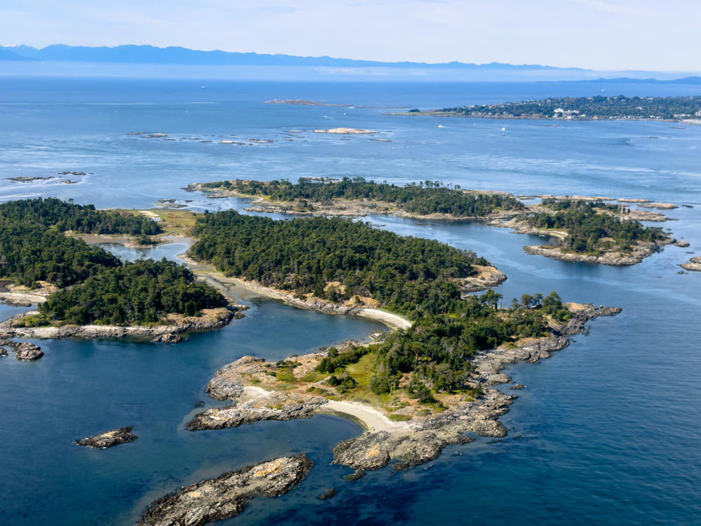 Aerial view of a cluster of small, forested rocky islands with dense evergreen trees, rugged shorelines, and pale gravel beaches, surrounded by blue ocean water. In the distance a coastal town and a hazy mountain range meet the horizon under a pale blue sky.