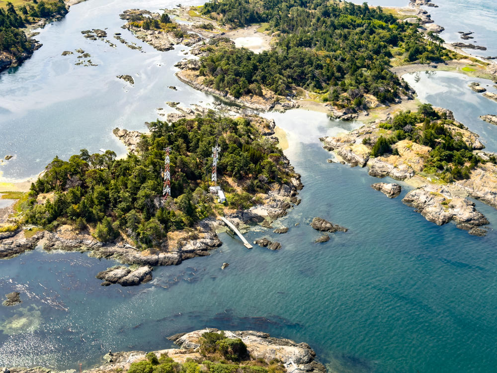 An aerial view of a cluster of small forested islands surrounded by calm blue-green water, rocky shorelines, and exposed tidal flats. Two red-and-white lattice communication towers rise above the trees on the central island, near a small building and a wooden dock reaching into the channel.