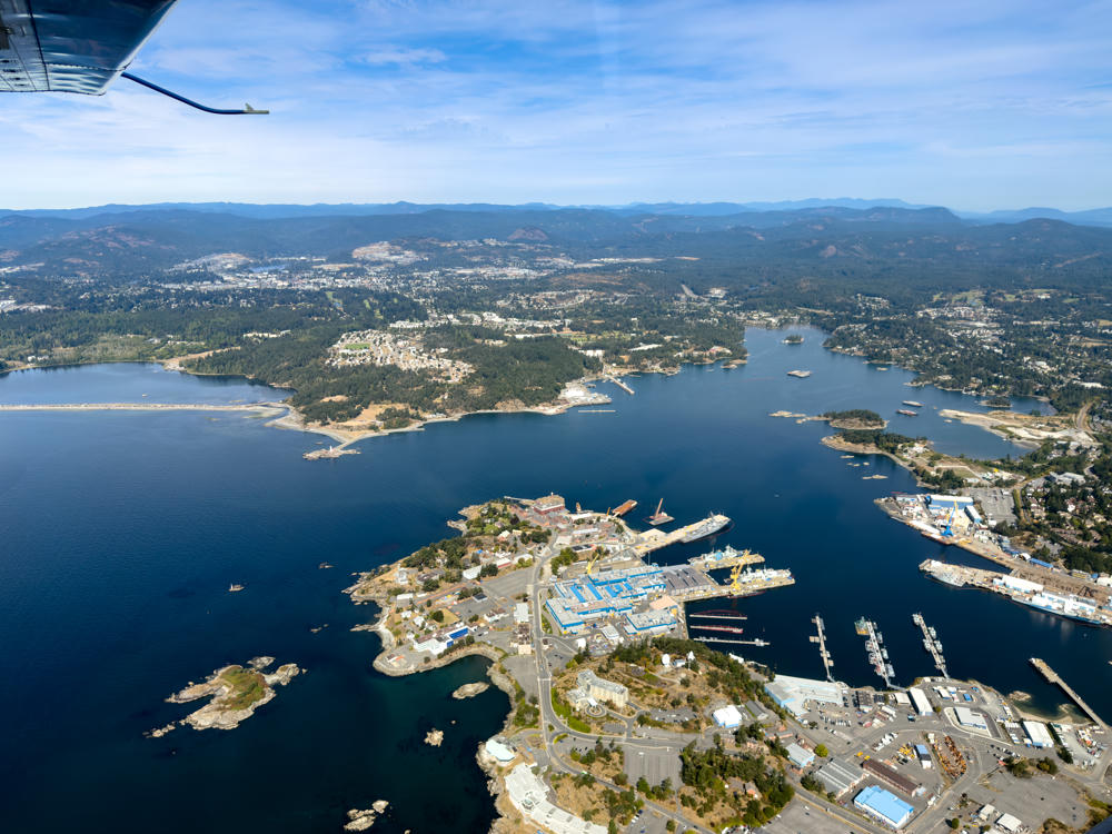 An aerial view from a small aircraft, with a wing strut and part of the plane visible in the upper left, looking down over a peninsula shipyard and naval harbour surrounded by dark blue water, with wooded suburbs and distant mountains under a hazy blue sky.