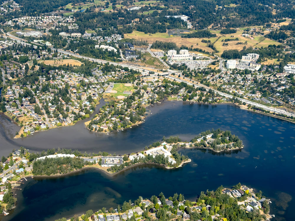 An aerial view over Saanich, British Columbia, showing a dark, meandering lake or inlet dotted with small wooded islands and peninsulas lined with houses, apartment buildings, and mature evergreens. In the distance a highway winds through suburban neighbourhoods, a baseball diamond and tennis courts, mid-rise buildings, and rolling forested hills and dry golden fields under bright daylight.