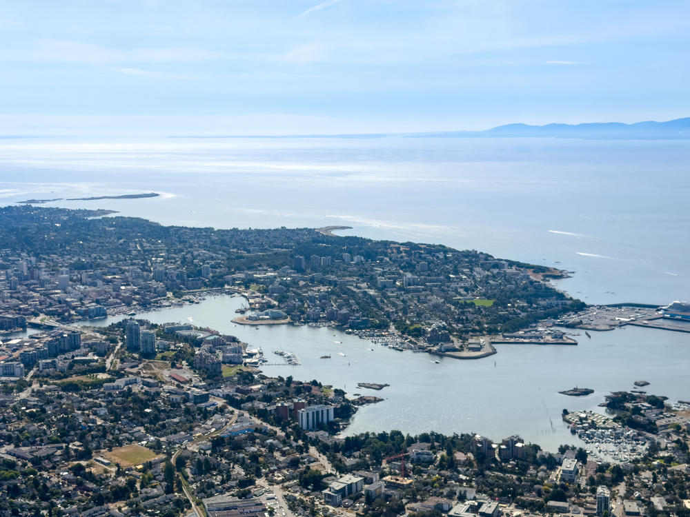 Aerial view over Esquimalt and Victoria, British Columbia, showing a sprawling coastal city of low buildings, marinas and harbours winding between wooded peninsulas, with the shimmering Strait of Juan de Fuca and hazy distant mountains beyond under a pale blue sky.