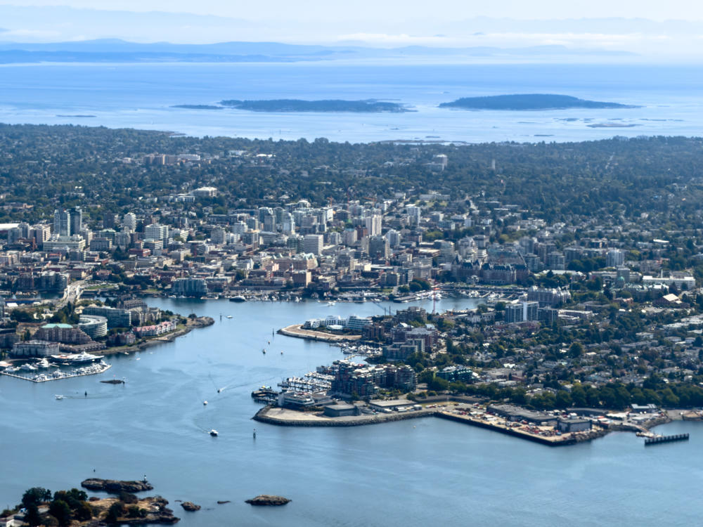 Aerial view of a coastal city built around a harbor, with clustered downtown high-rises, marinas full of moored boats, and residential neighborhoods spreading into forested hills, all fronting the broad blue waters of the Salish Sea with low islands and hazy distant mountains on the horizon.