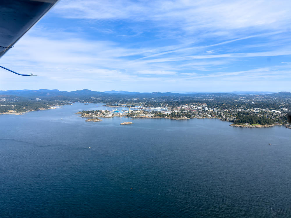 An aerial view from a small aircraft, with a wing strut and float visible in the upper left, looking over the blue waters of the Salish Sea toward a rocky, wooded coastline and harbour with docks, cranes, and buildings near Esquimalt, backed by low forested hills under a bright sky with wispy clouds.
