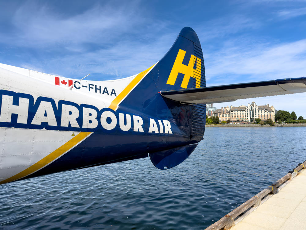 The rear fuselage and tail of a Harbour Air seaplane with registration C-FHAA, painted in blue, white and yellow with a Canadian flag, floating on the calm water of Victoria Harbour with a waterfront dock and hotel buildings across the water under a partly cloudy blue sky.