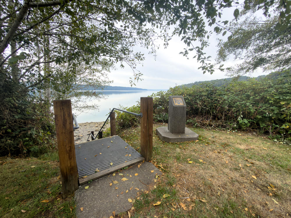 A wooden boardwalk with metal handrails leads down toward a calm inlet at Lilliwaup Tidelands State Park, framed by overhanging tree branches. A concrete pedestal with a bronze plaque sits on the grass to the right, with forested hills and an overcast sky across the water.