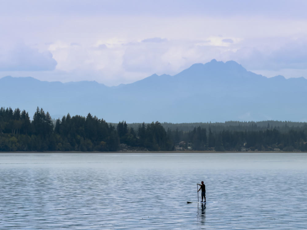 A lone silhouetted figure stands on a paddleboard on calm gray water, with a forested shoreline and dwellings across the way and hazy layered mountains rising into a soft overcast sky.