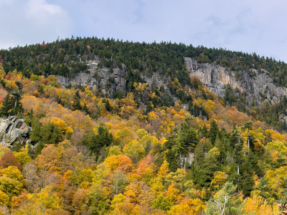 A forested mountainside ablaze with autumn color in oranges, reds, yellows and greens, topped by exposed gray granite cliffs and evergreen trees along the ridgeline under an overcast pale sky.