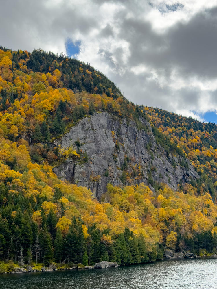 A rugged gray rock cliff rising above a mountainside covered in autumn foliage of orange, yellow, and green, with evergreens along the shoreline of a dark lake in the foreground under a cloudy sky.