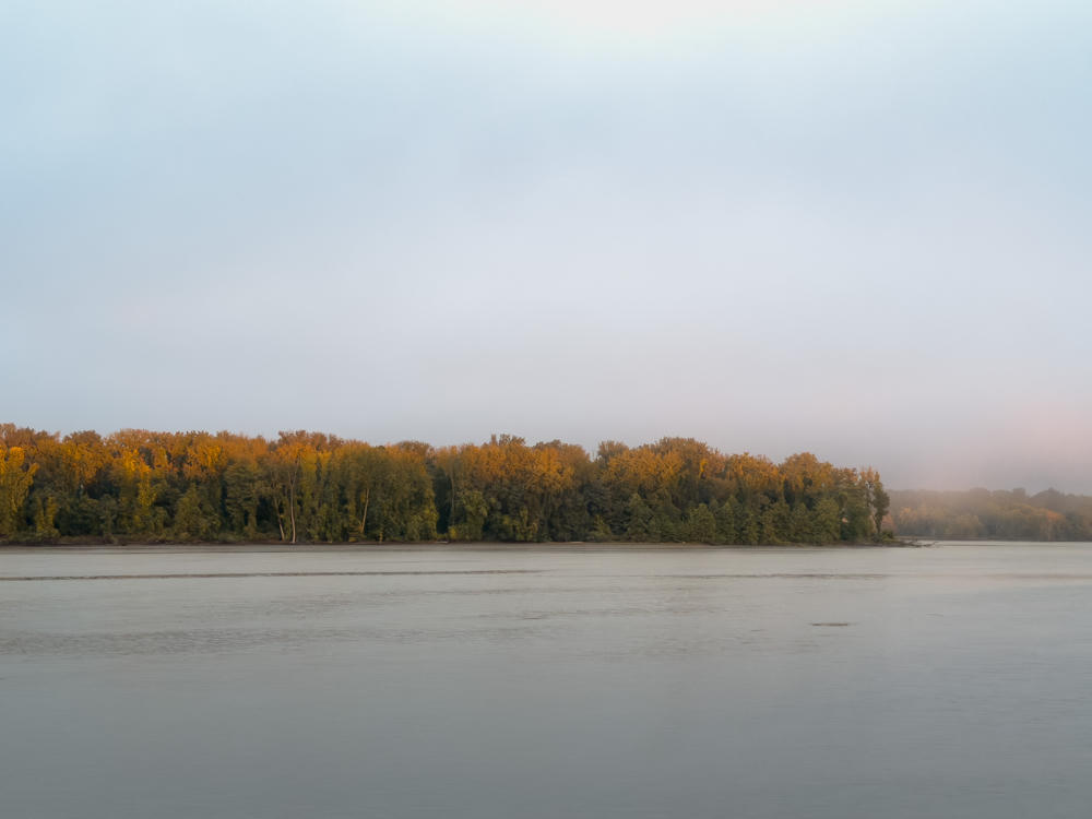 A wide, calm river stretches across the foreground beneath a hazy, pale gray sky, with a dense line of trees along the far bank catching warm golden morning light. Mist softens the far shoreline on the right, and the autumn foliage glows orange and yellow above darker green growth.