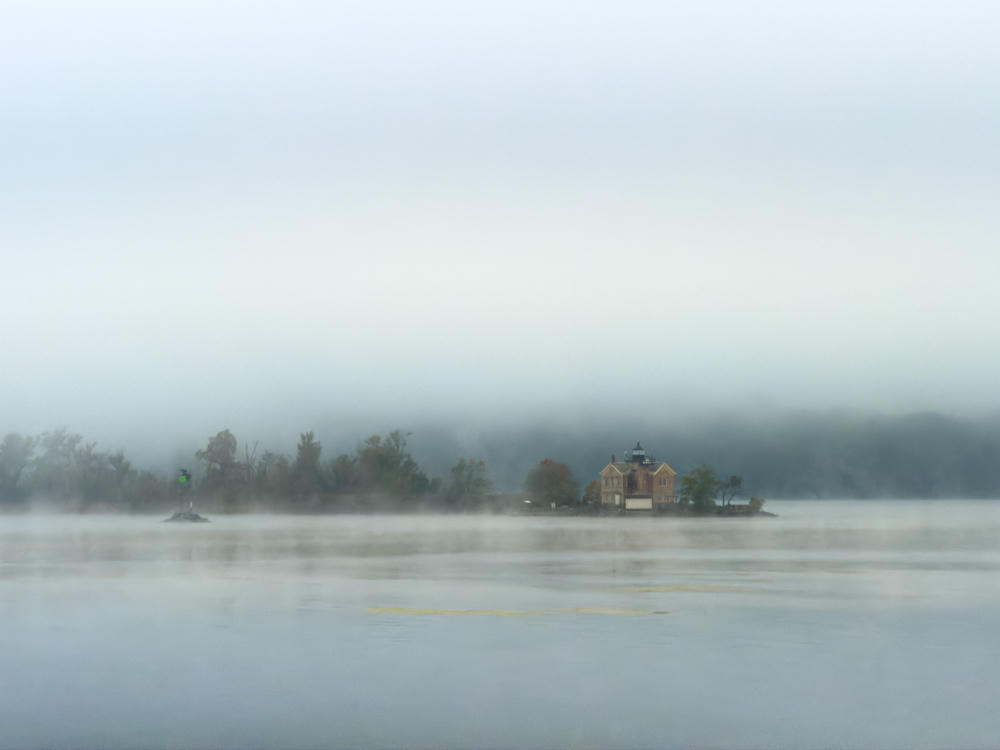 A brick lighthouse building and a green navigation marker rise from the misty water of a river at dawn, with a wooded shoreline partly obscured by heavy fog and low cloud filling the pale sky.