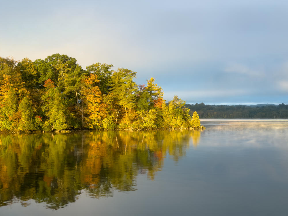 A calm bay in early morning light with a wooded shoreline of trees in fall colors — green, gold, and orange — mirrored in the still water, under a hazy sky with low clouds; a faint reflection on a window suggests the shot was taken from indoors.