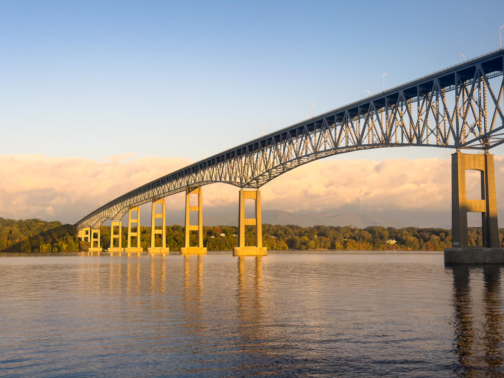 A long steel truss bridge spans a wide river in warm early-morning light, its concrete piers reflecting on the calm water. Low clouds hang over tree-lined banks with hints of autumn color and distant hills beyond.