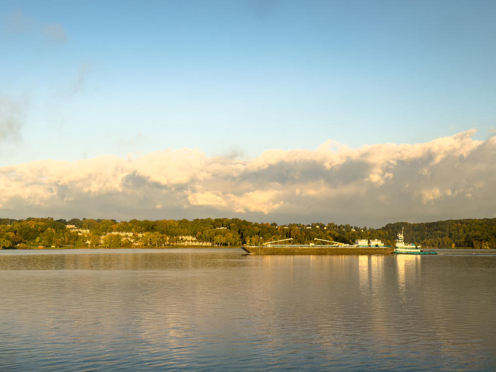 A tugboat pushing a long cargo barge along a broad, calm river in warm early morning light, with a tree-lined far shore dotted with buildings and a bank of low clouds beneath a pale blue sky.