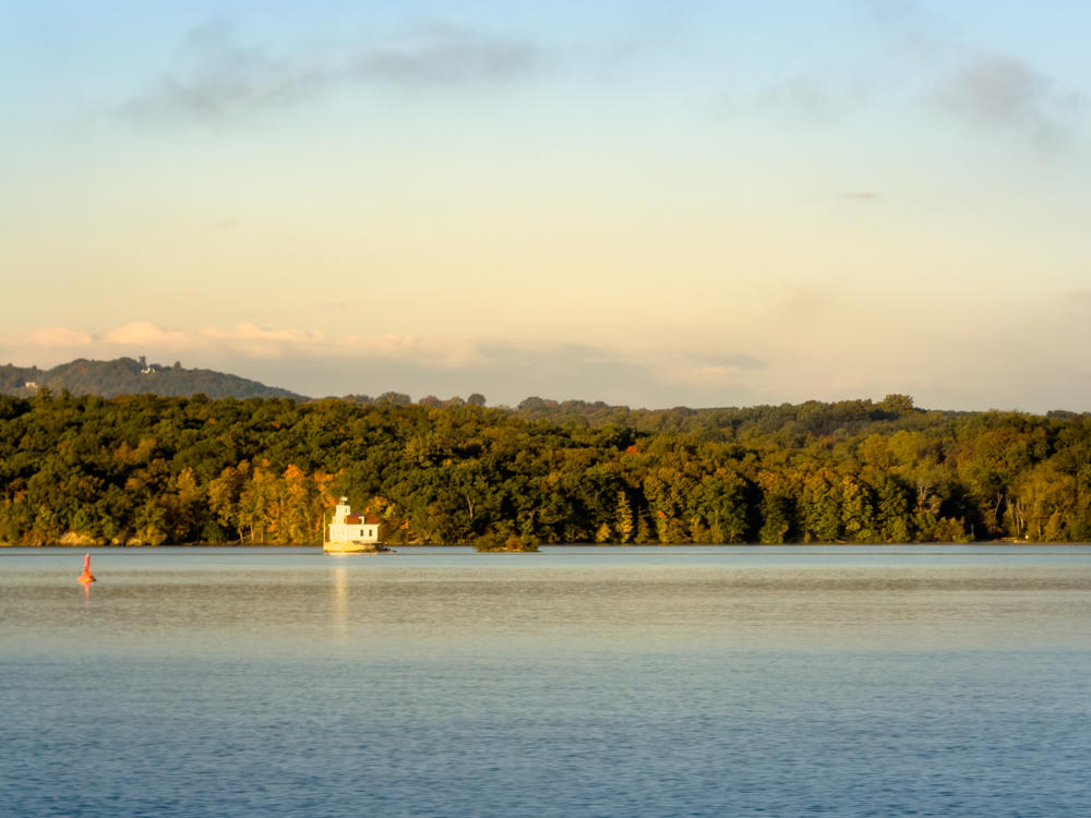 A calm, wide river in early morning light, with a small white lighthouse on a wooded shoreline and a red buoy floating in the water. Low tree-covered hills line the far bank beneath a pale sky with soft clouds.