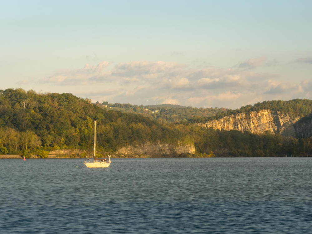 A sailboat with its mast bare sits moored on calm river water in the morning light, with a forested shoreline and exposed pale rock cliffs rising beyond, under a soft hazy sky with scattered clouds.