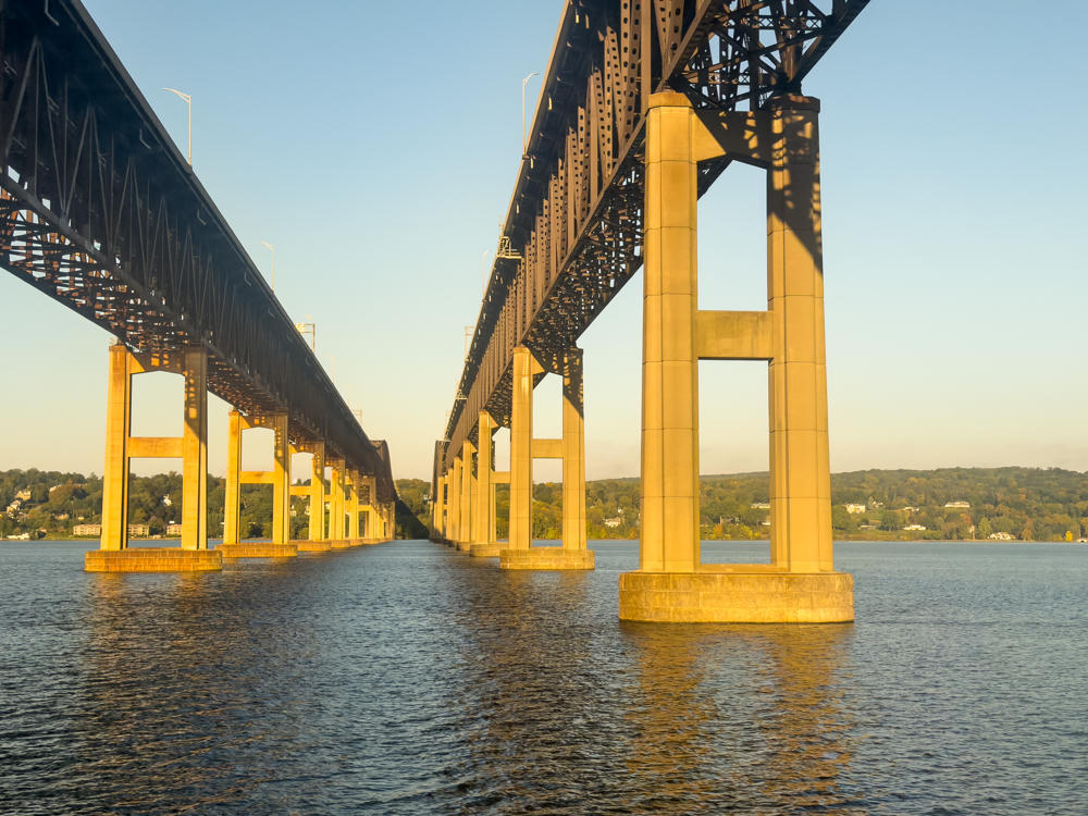 A view from water level beneath two parallel steel truss bridges spanning a wide river, their tall concrete piers rising from the water and lit warmly by low morning sun against a clear blue sky, with wooded hills and shoreline buildings in the distance.