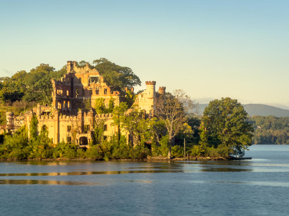 The ruins of Bannerman Castle on Pollepel Island in the Hudson River, its crumbling brick walls, turret, and overgrown vegetation glowing in warm early-morning light, with calm water in the foreground and wooded hills along the far shore under a pale clear sky.