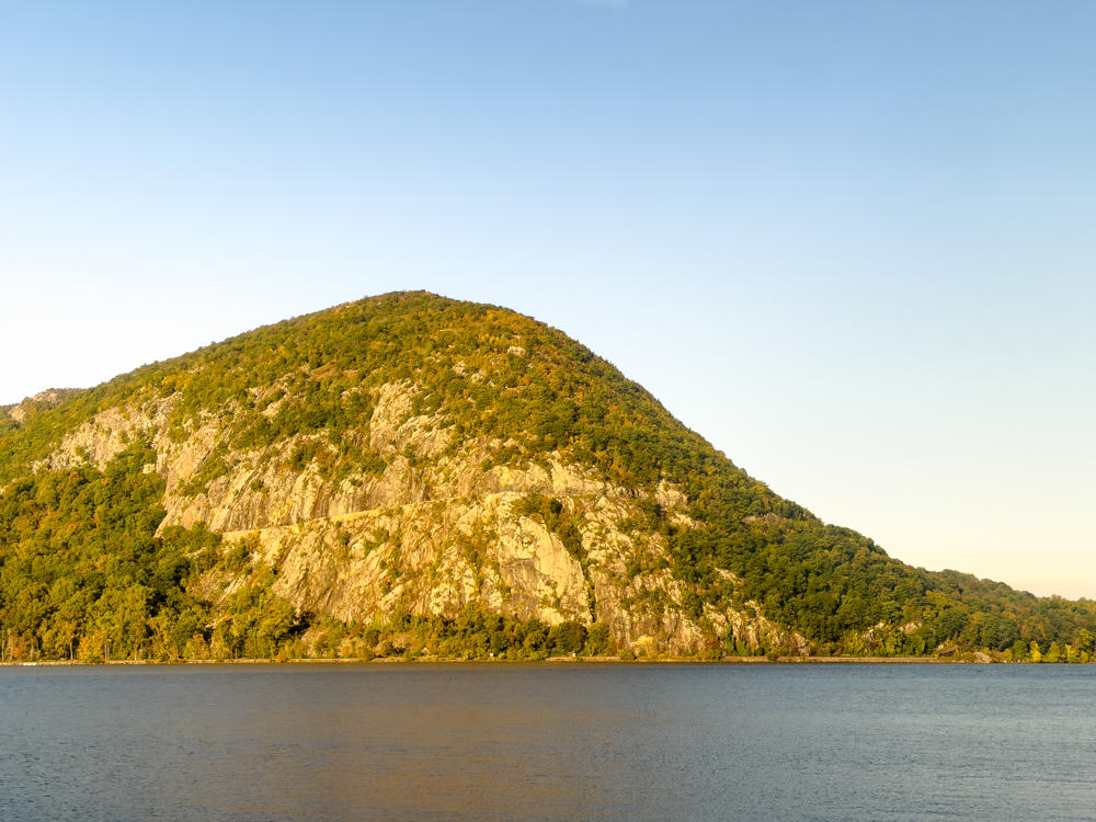 A rounded, tree-covered mountain with a prominent bare granite cliff face rising above the far bank of a wide, calm river under a clear pale blue morning sky. Warm early light catches the rock and foliage, with hints of autumn color in the trees along the shoreline.