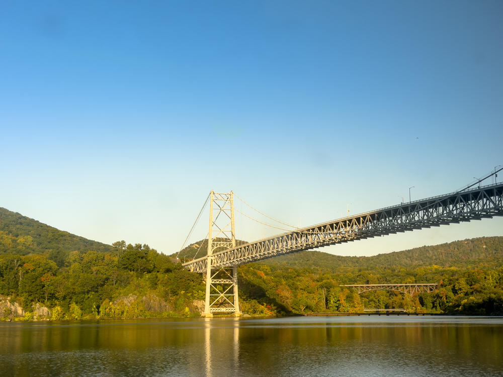 A steel suspension bridge spans a wide river, with its tall tower and lattice cables lit by warm morning light, backdrop of forested hills showing early autumn color under a clear blue sky. A smaller steel truss railway bridge sits lower along the far shoreline.