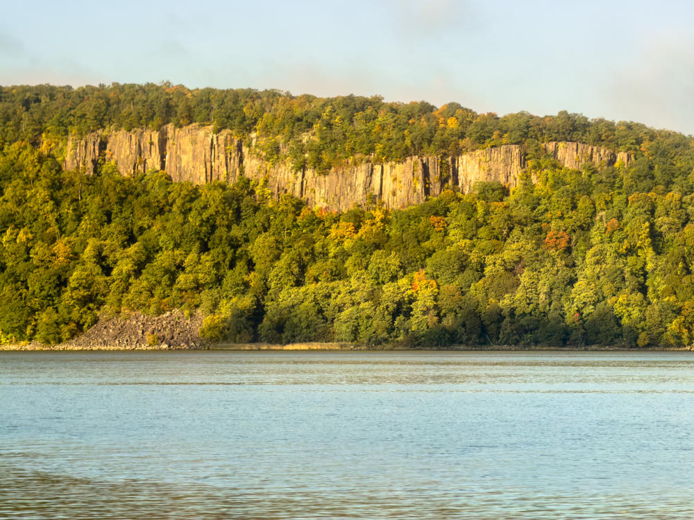 A view across the Hudson River toward the steep columnar basalt cliffs of the Palisades, rising above a dense forested slope just beginning to show autumn color under soft morning light. A hazy pale blue sky stretches above the ridgeline, with a bank of low cloud or mist on the right.