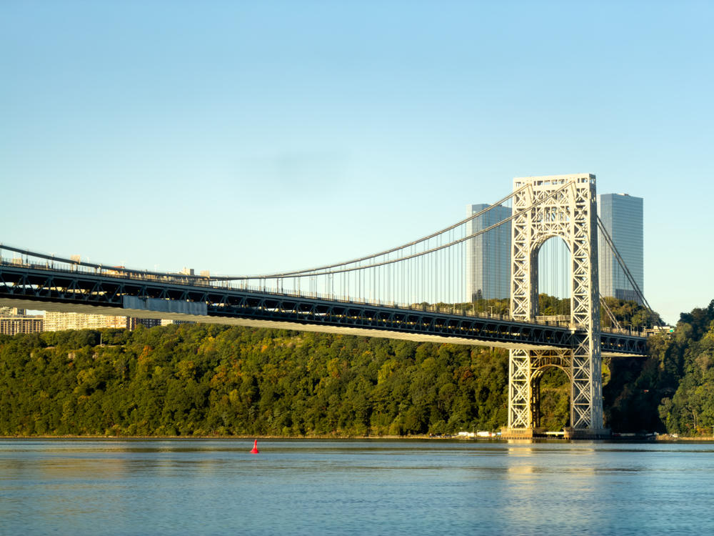 The George Washington Bridge, a steel suspension bridge, spans the Hudson River under a clear blue morning sky, with a tree-covered cliff and tall buildings on the far bank and a red buoy floating on the calm water in the foreground.