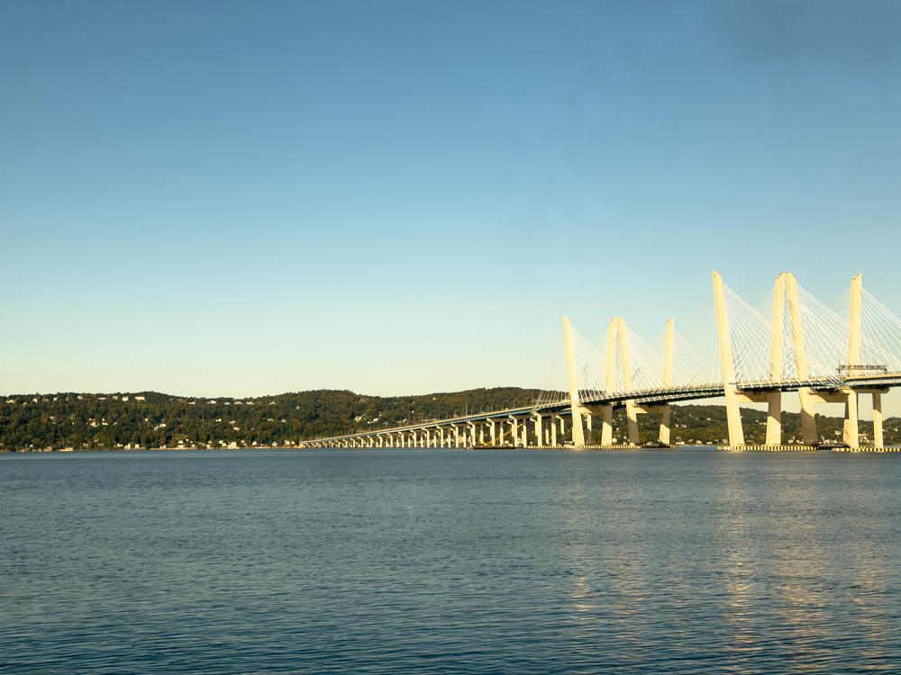 A long cable-stayed bridge with tall white towers spans a wide river under a clear blue sky, with low forested hills lining the far shore in warm morning light.
