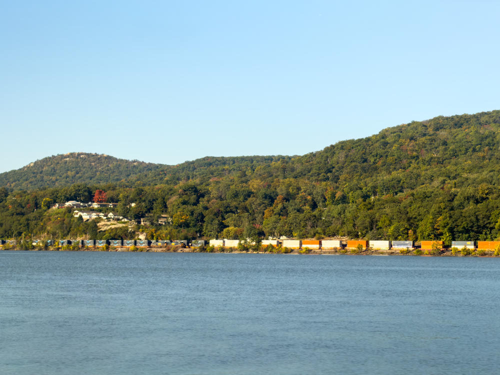 A long freight train with colorful intermodal containers and freight cars runs along the far shore of a wide river, below forested hills touched with early autumn color. Clusters of houses sit on a rocky bluff above the tracks under a pale, clear sky.