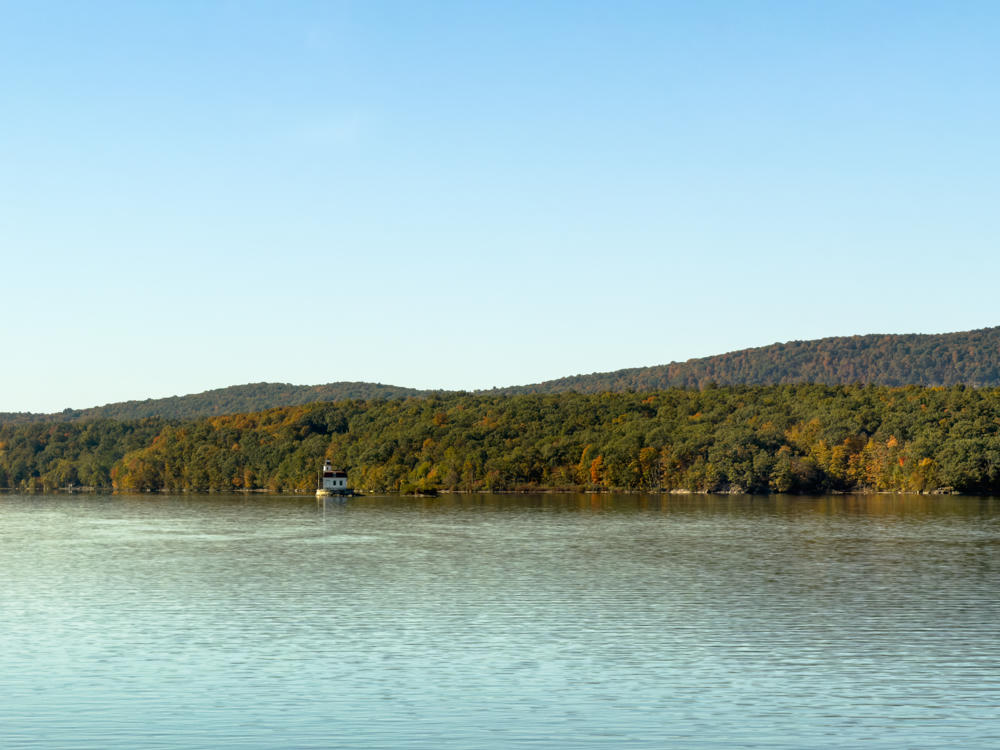 A calm, wide river stretches across the foreground beneath a pale blue sky, with a small white lighthouse sitting at the water’s edge in front of a forested hillside showing early autumn colors of green, gold, and orange.