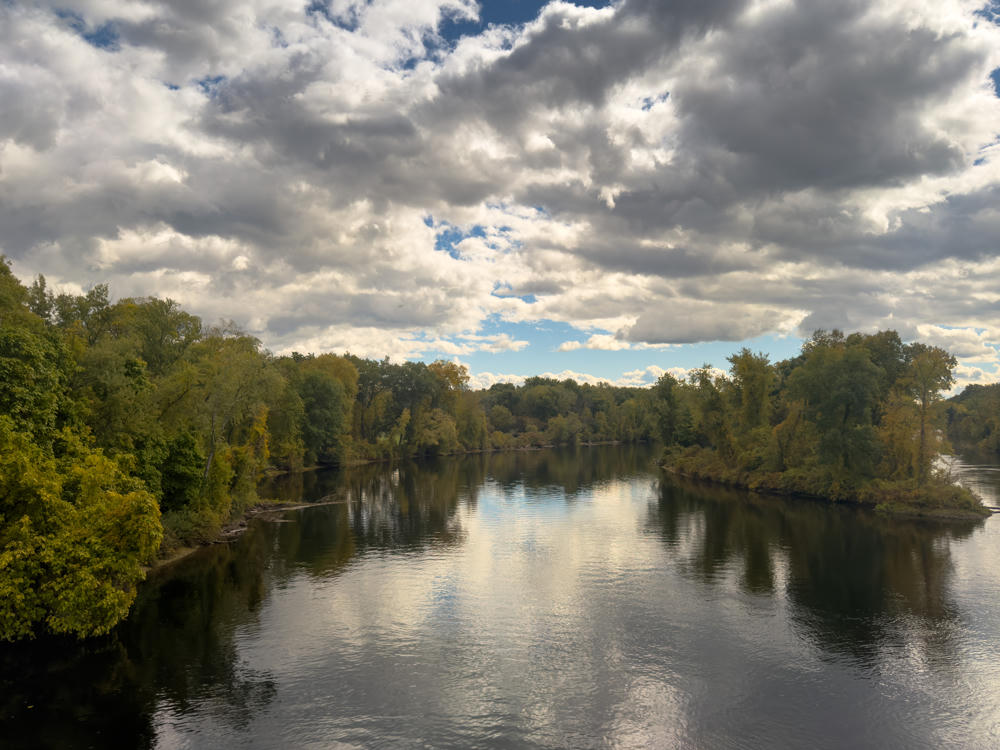 A calm, wide river lined with dense green trees just beginning to turn autumn colors, under a dramatic sky of heavy white and gray clouds with patches of blue. The still water reflects the clouds and surrounding foliage.