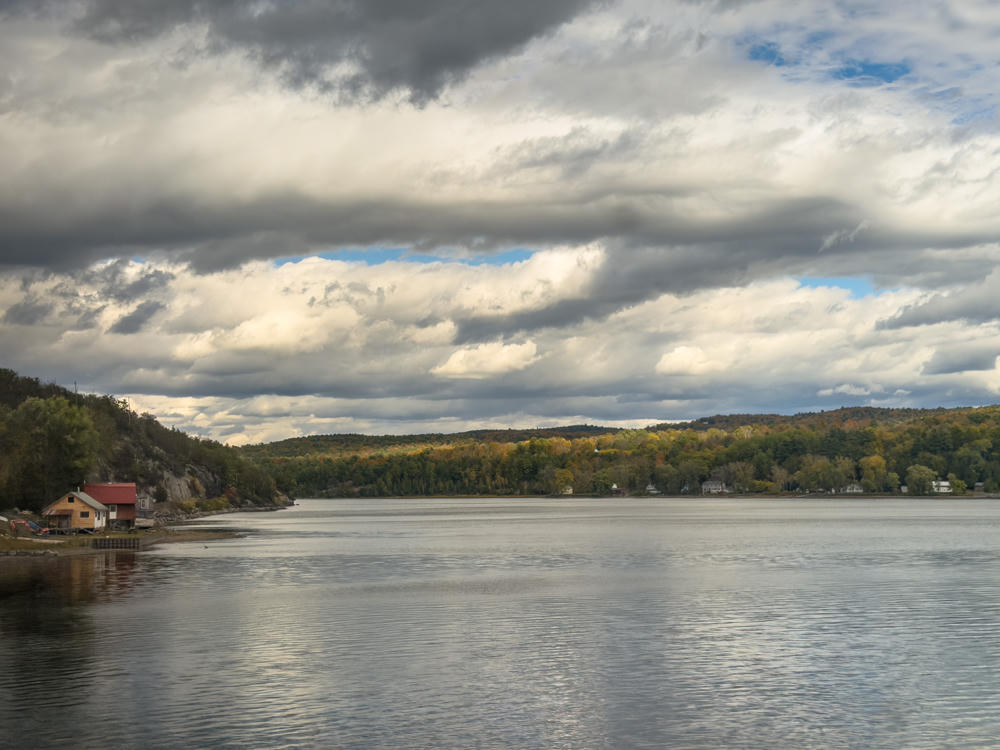 A broad, calm lake under a dramatic sky filled with heavy gray and white clouds broken by patches of blue. Wooded hills lined with early autumn foliage run along the far shore, while a small cabin with a red roof and a parked excavator sit on a rocky point at the left edge of the water.