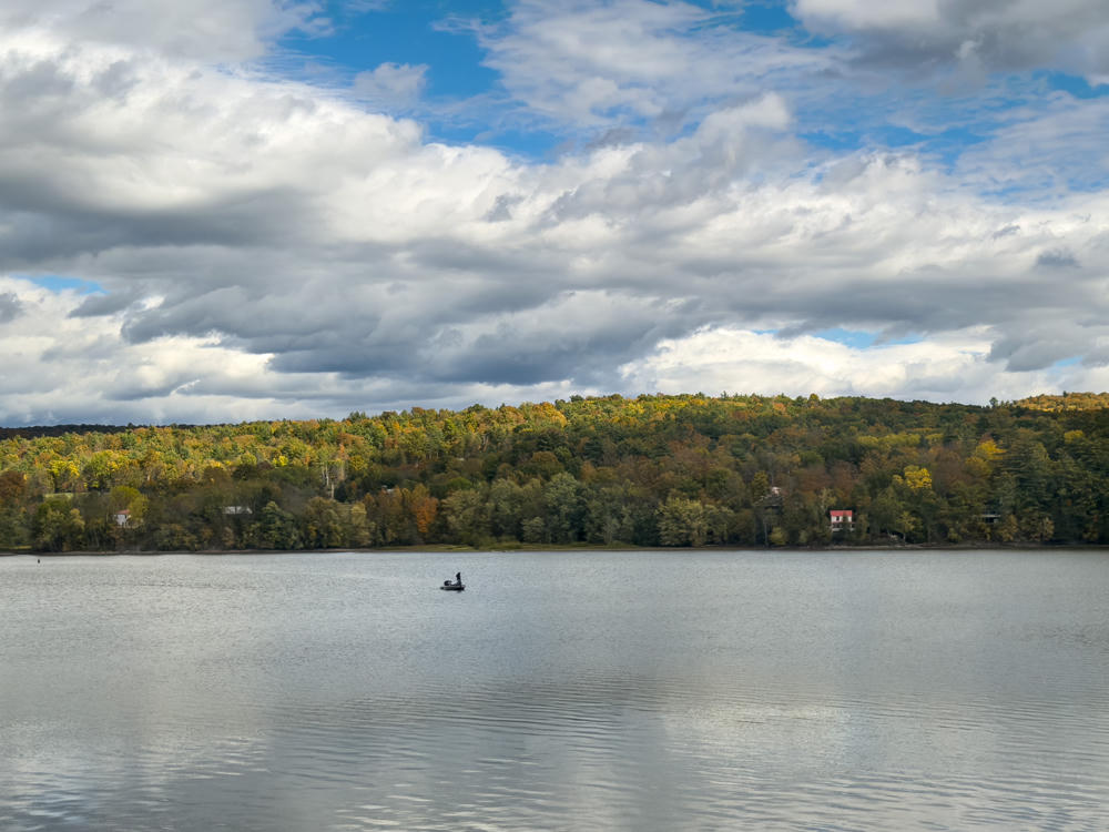 A lone person in a small boat sits on a calm lake beneath a dramatic sky of broken clouds, with a far shore of hills covered in autumn-colored forest and a few scattered houses along the waterline.