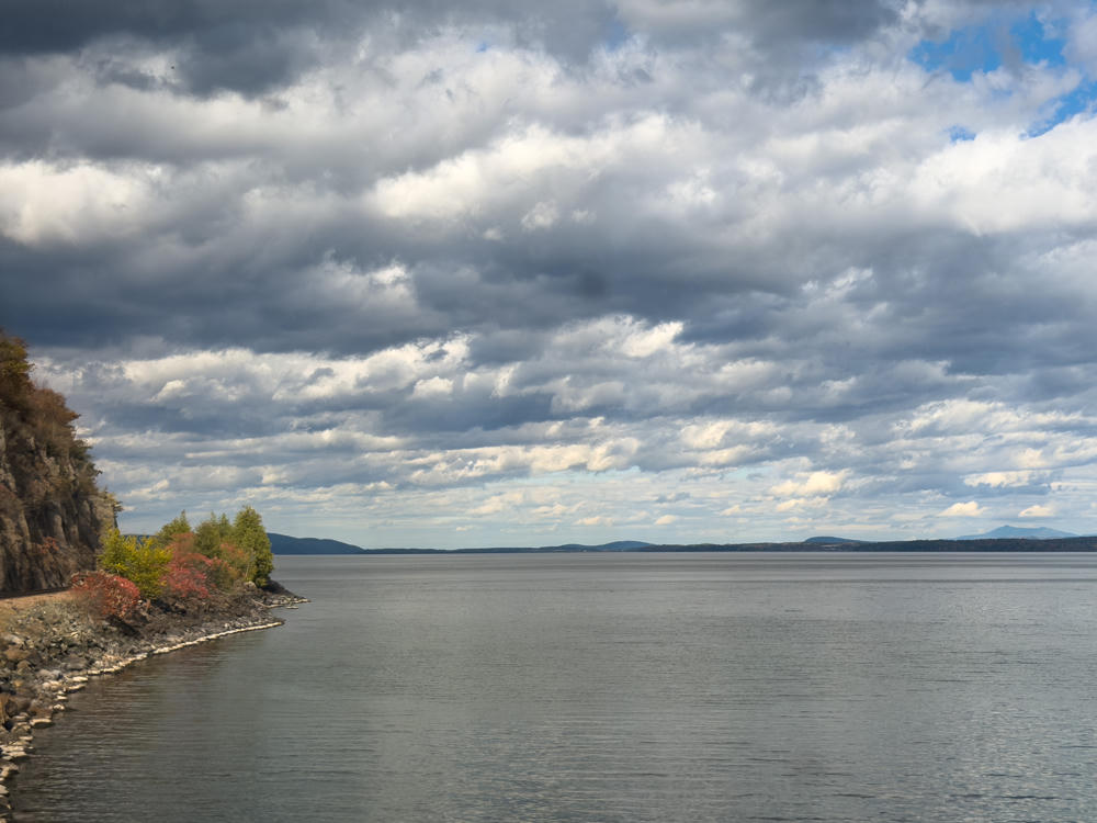 A broad lake under a dramatic, overcast sky filled with layered gray and white clouds, with distant low mountains along the far shore. In the left foreground a rocky cliff and shoreline are dotted with trees showing early autumn color.