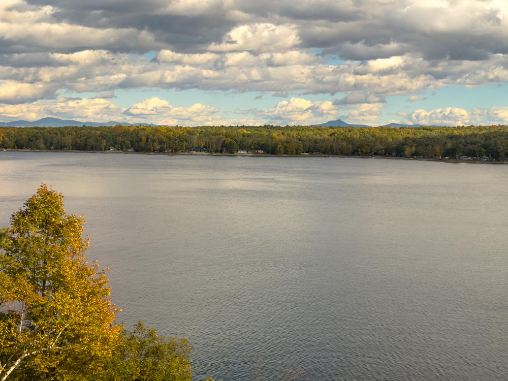A broad view across a calm lake under a dramatic sky of scattered white and gray clouds, with a forested far shore dotted with a few houses and distant blue mountain ridges on the horizon. In the foreground, trees with autumn-tinged yellow and green foliage rise from the near shore.